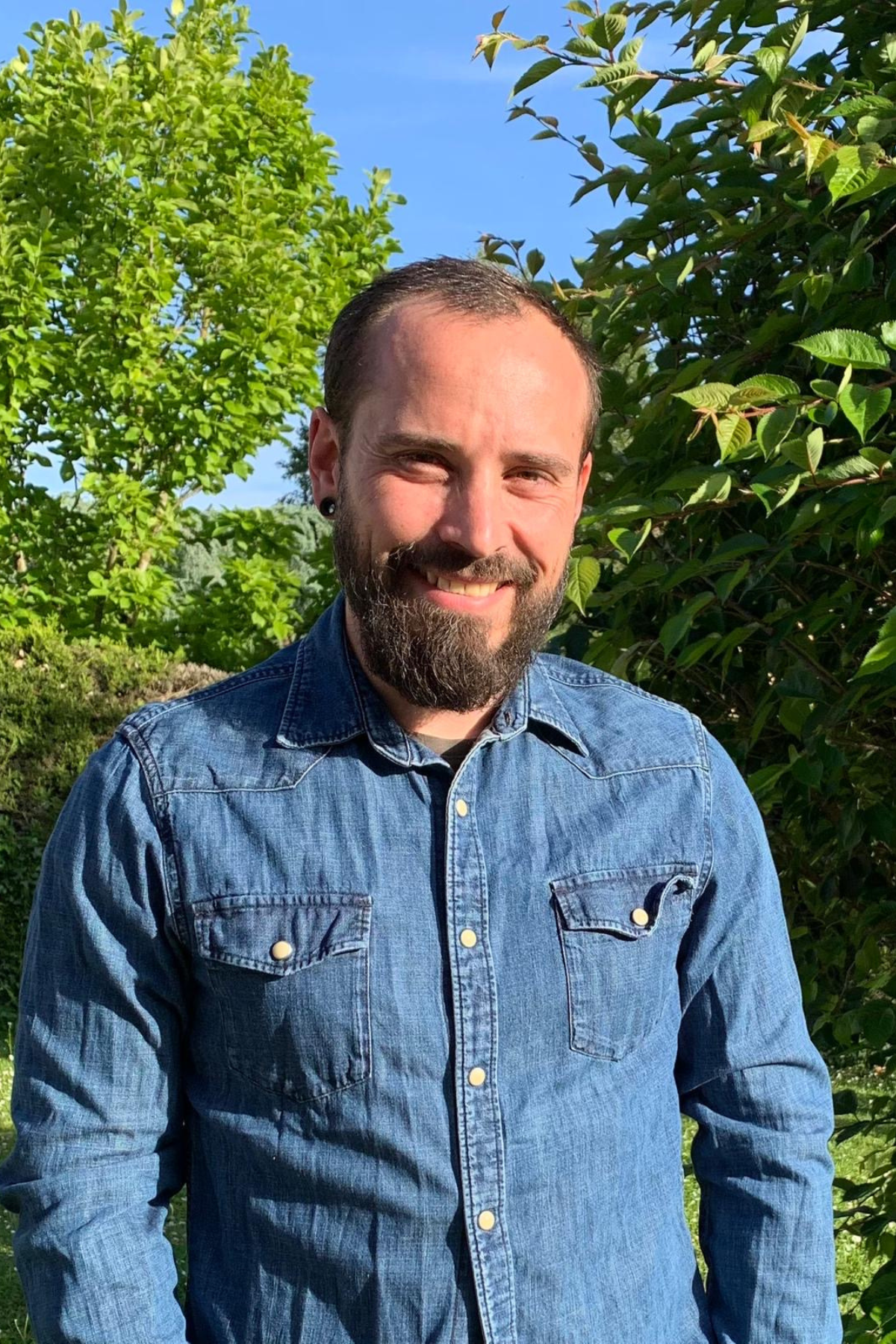 Homme souriant portant une chemise en jean, debout à l'extérieur par une journée ensoleillée. Il se tient devant une végétation dense avec des arbres et des buissons verts en arrière-plan.